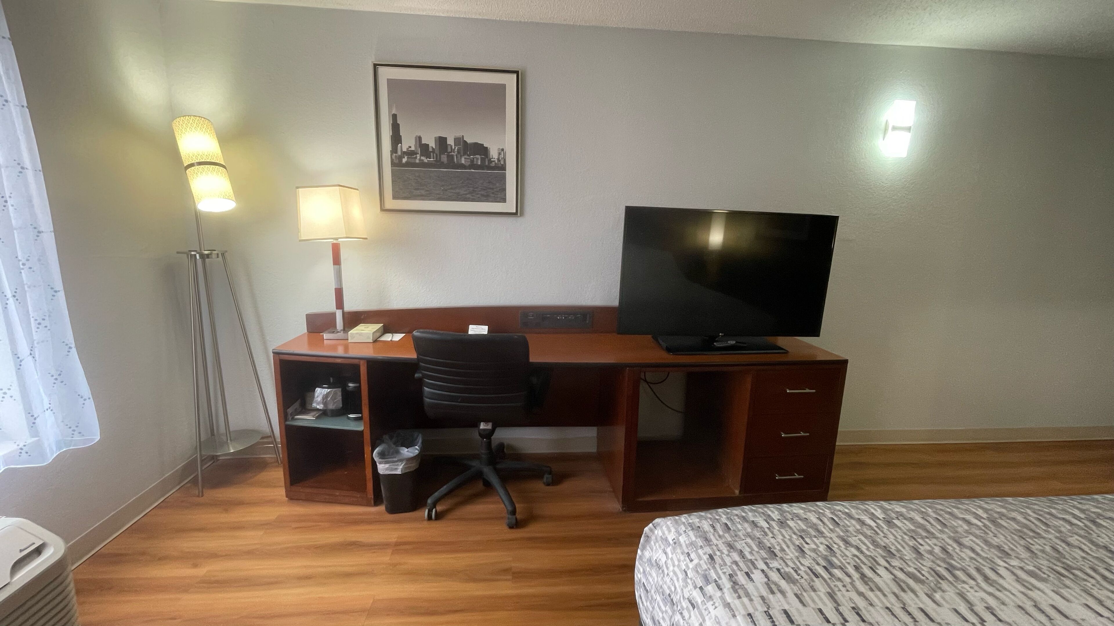 Habitación de hotel con un escritorio de madera, una silla de oficina negra, un televisor, un cuadro en el pared con una vista urbana enmarcada y una cama con una colcha estampada.