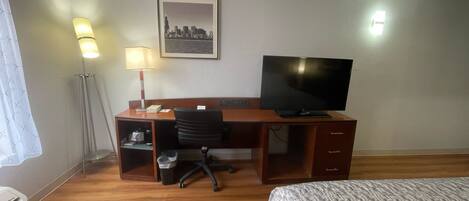 Habitación de hotel con un escritorio de madera, una silla de oficina negra, un televisor, un cuadro en el pared con una vista urbana enmarcada y una cama con una colcha estampada.
