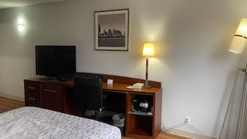 Ein Hotelzimmer mit einem Bett, einem Schreibtisch mit Fernseher, einem Sessel, einem Stadtbild im Bilderrahmen, einer Lampe und einem Mülleimer.