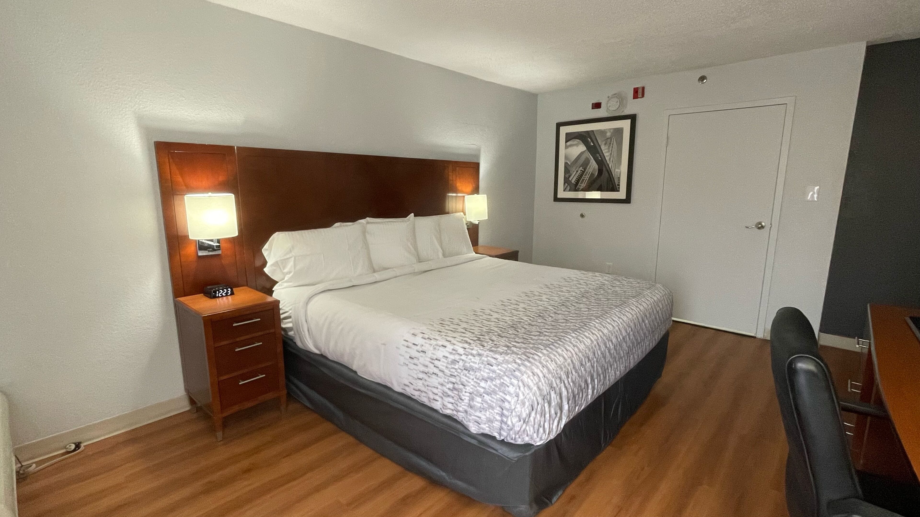 Une chambre d’hôtel avec un grand lit, une tête de lit en bois, deux tables de chevet avec des lampes, un tableau encadré au mur et une porte.