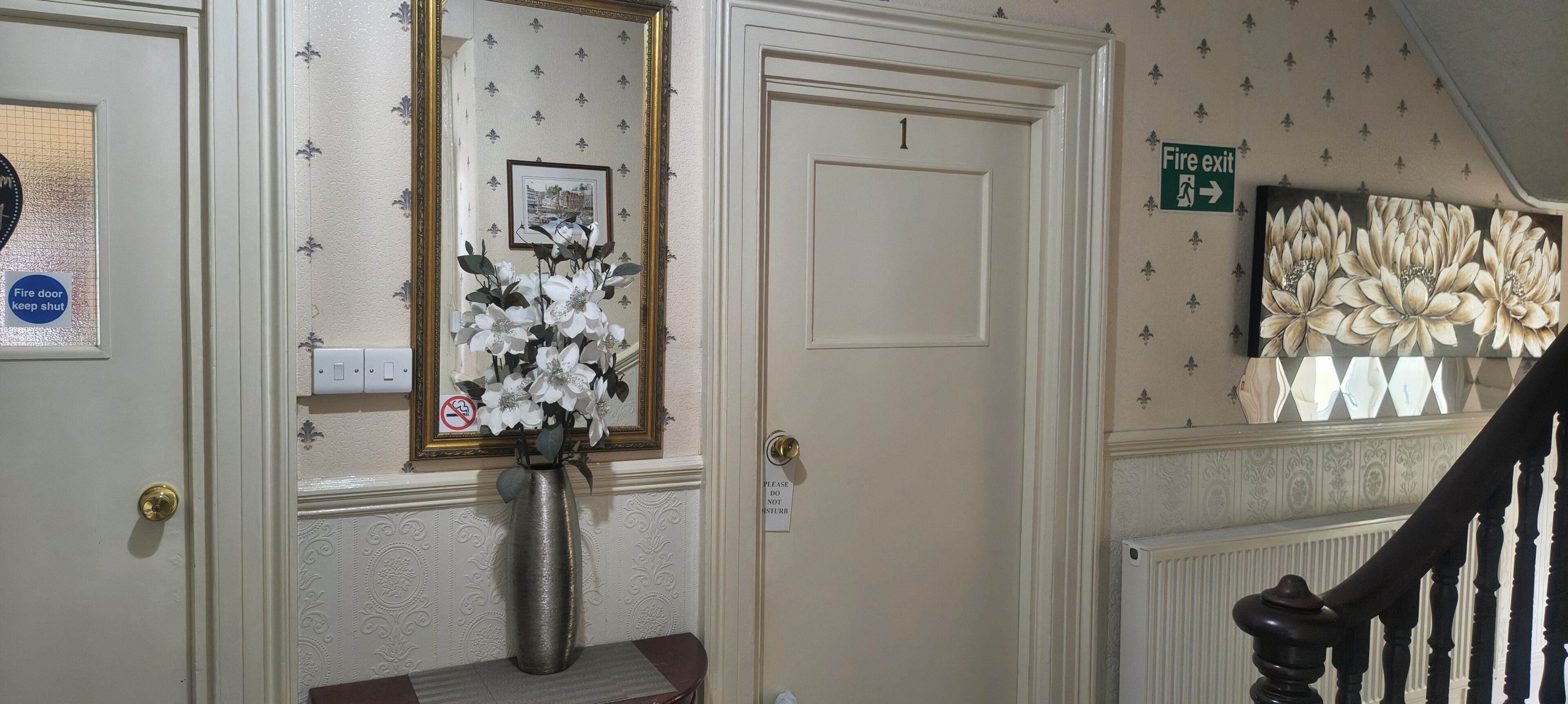 A hallway with a white door, a framed picture, a vase with flowers, a 'Fire door keep shut' sign, and a 'Fire exit' sign.