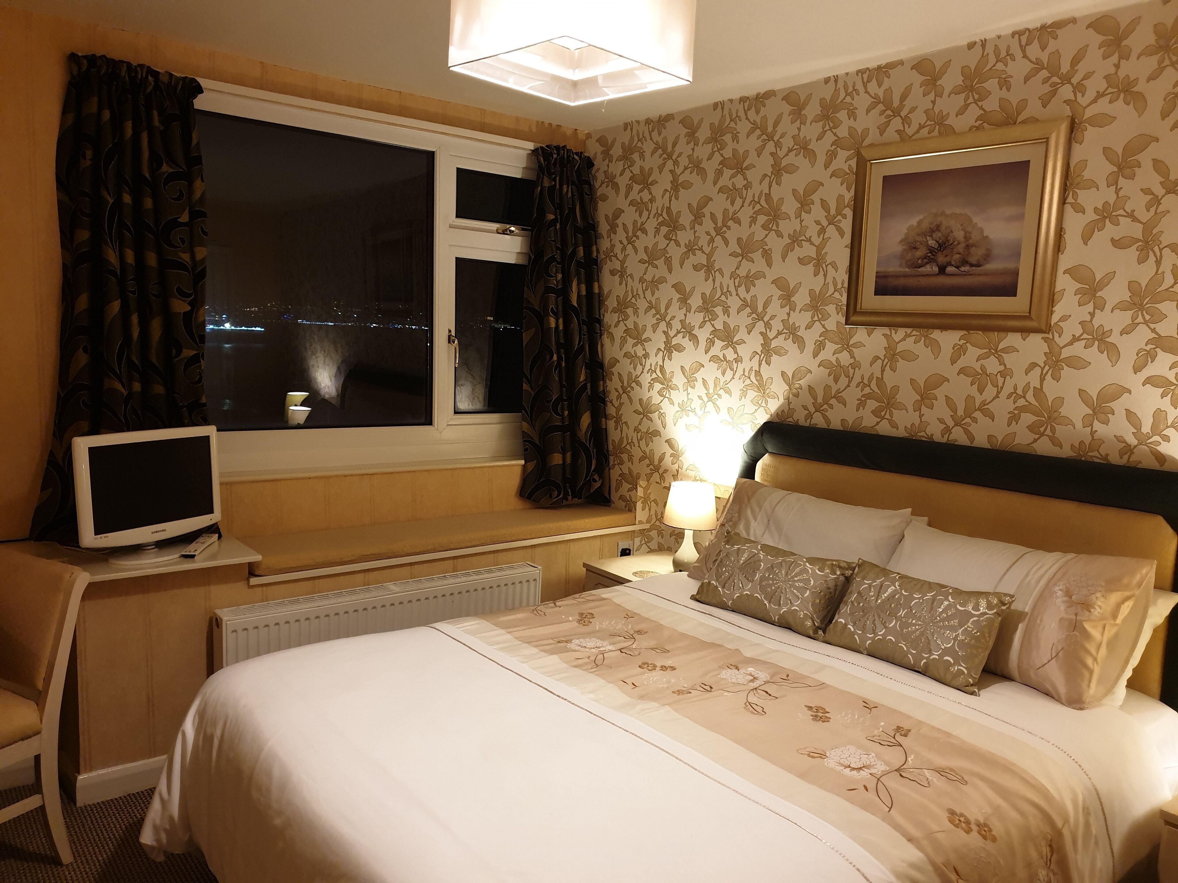 A hotel room with a bed, a desk with a computer, a window with curtains, a radiator, and a wall with a floral pattern.