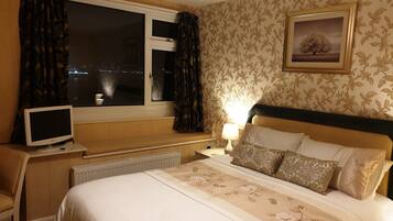 A hotel room with a bed, a desk with a computer, a window with curtains, a radiator, and a wall with a floral pattern.