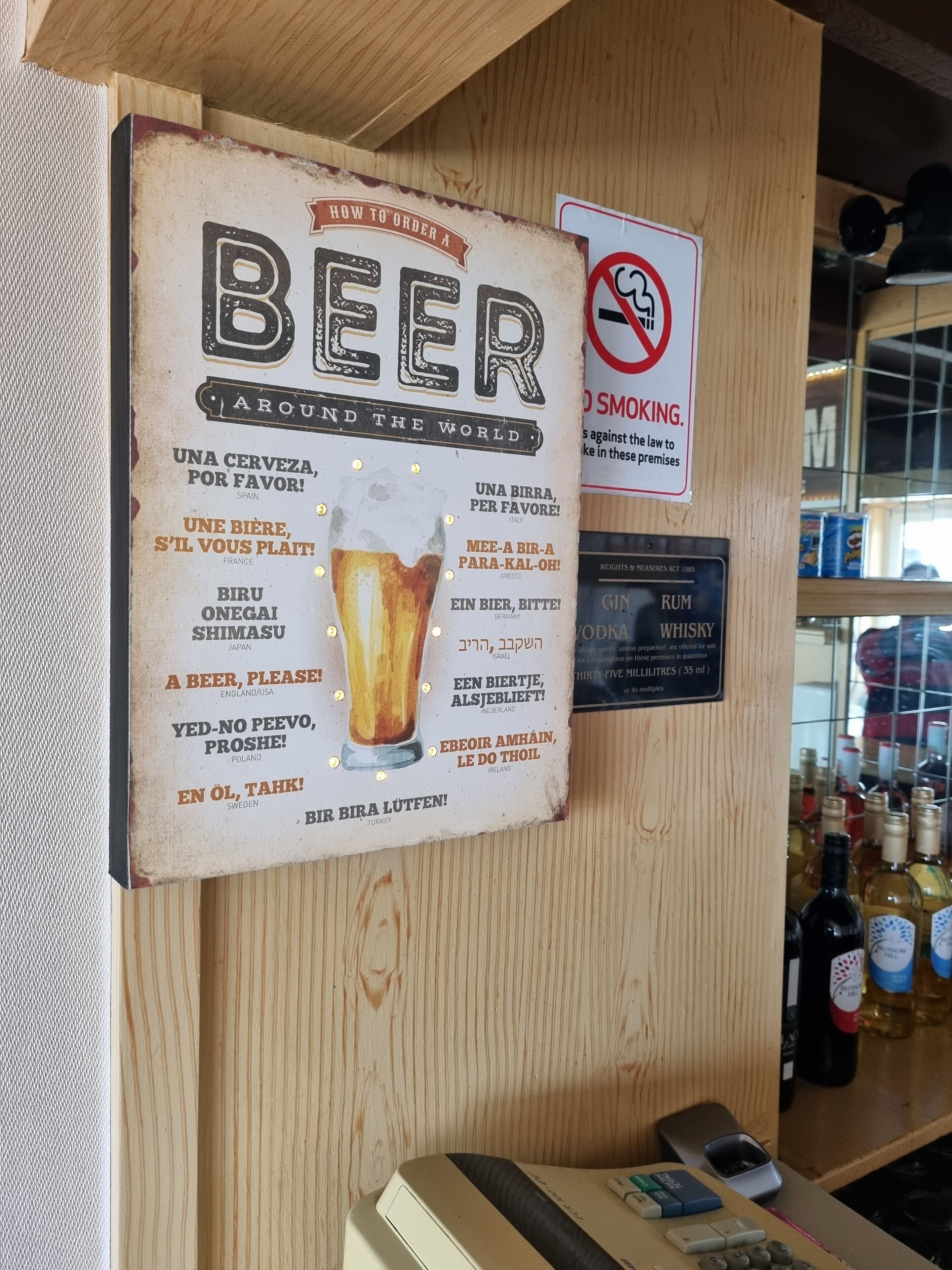Un mur sur lequel est affiché un panneau indiquant différents noms de bières dans différentes langues, un panneau interdisant de fumer et un autre indiquant les prix des boissons alcoolisées.