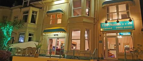 A hotel with a sign reading "The Sands Licensed" and a phone number. There are steps leading to the entrance, and the building has multiple windows and balconies.