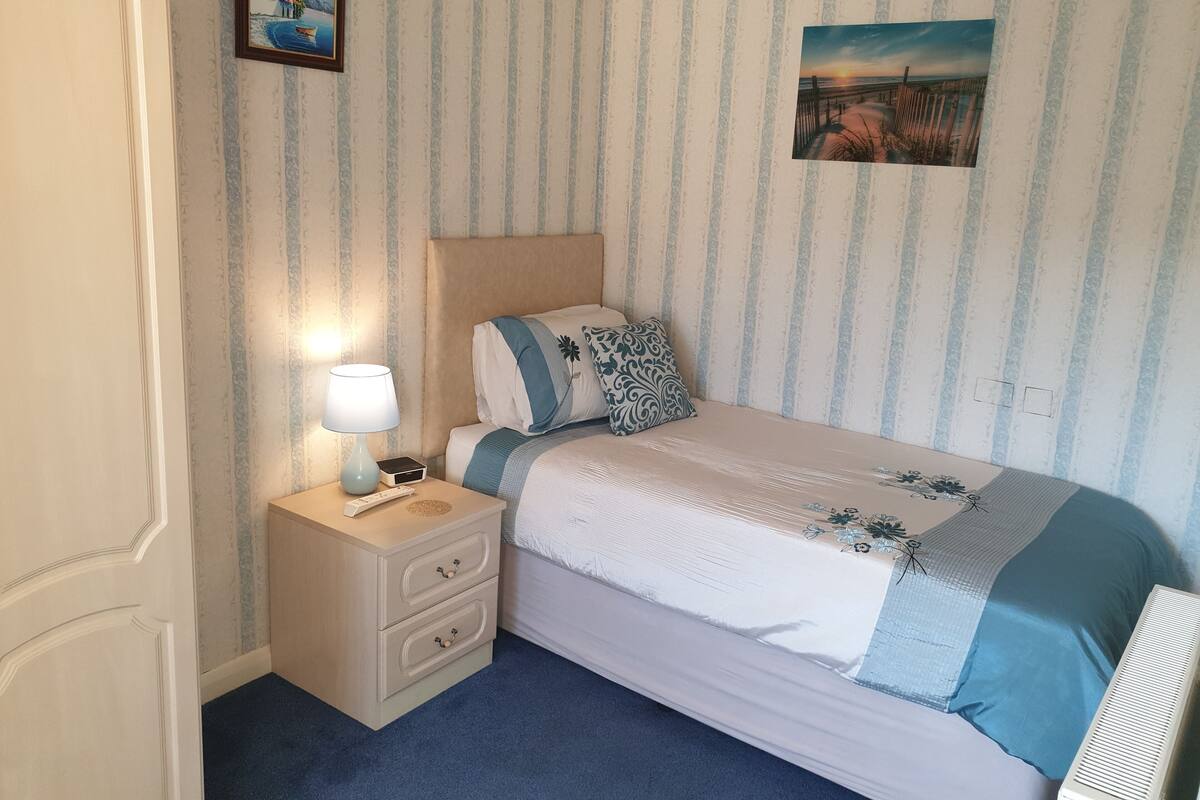 A bedroom with a bed, a nightstand, a lamp, a picture on the wall, and a power outlet.