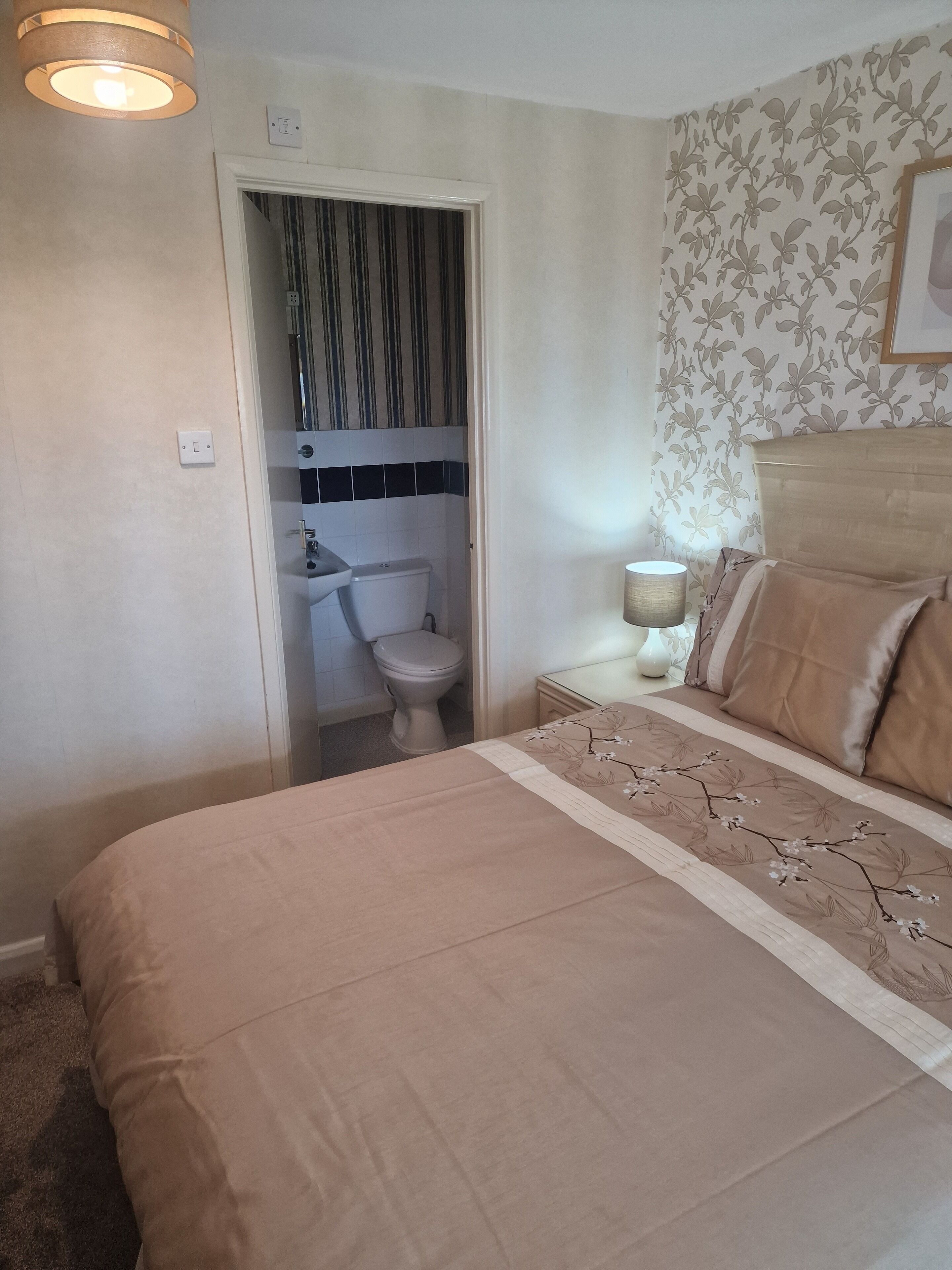 A bedroom with a bed, a bedside table with a lamp, a nightstand, a wall-mounted shelf, and a door leading to a bathroom.