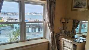 A room with a large window overlooking a coastal area, a desk with a lamp, and a dresser with drawers. - The Sands Hotel (Paignton)