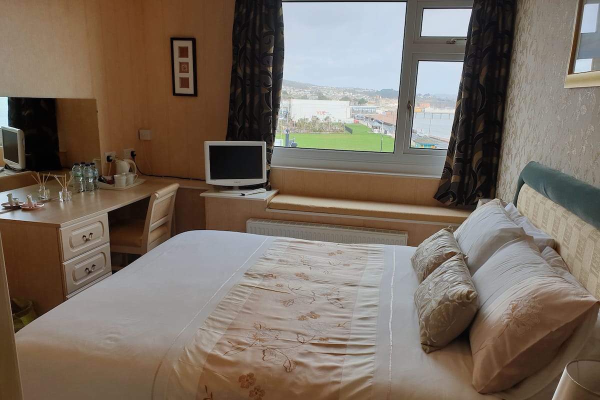 A hotel room with a bed, a desk with a computer, a window with a view of a cityscape, and a radiator.