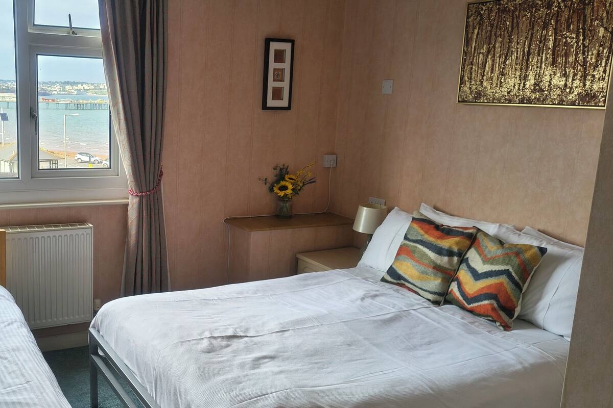 A hotel room with two beds, a window with a view of a bridge and water, a radiator, a nightstand with a vase of flowers, and a framed picture on the wall.