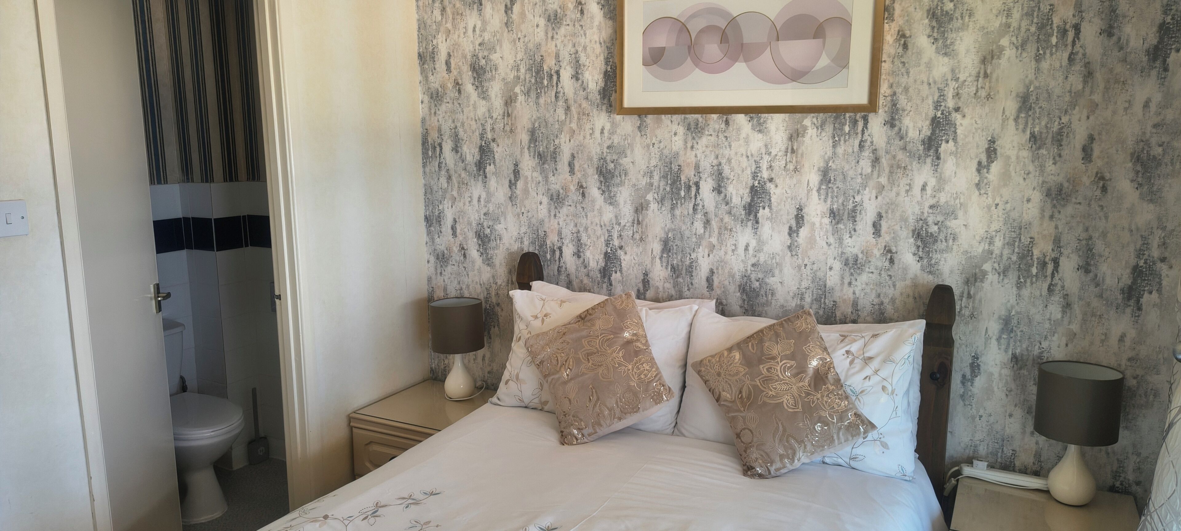 Une chambre à coucher avec un lit, deux tables de chevet, une commode, une œuvre d’art encadrée et une salle de bain visible à travers une porte ouverte.
