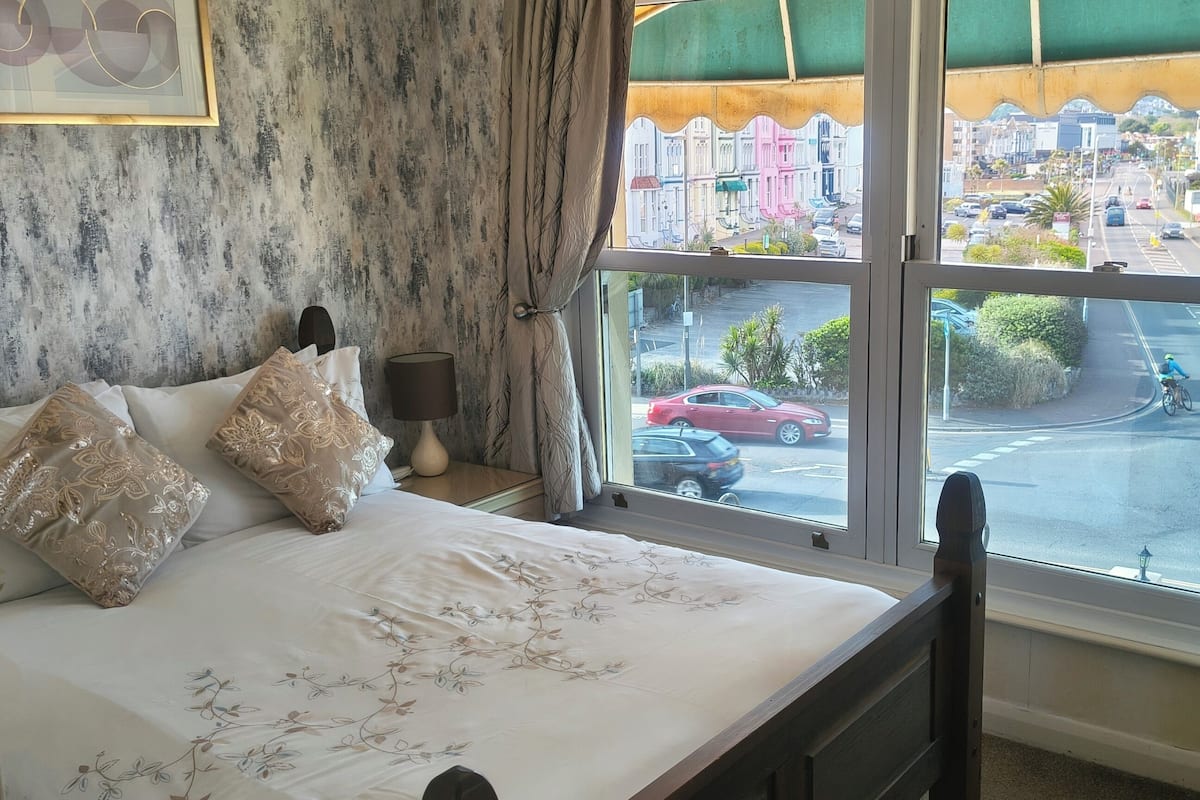 A bedroom with a bed, pillows, a lamp, and a view of a street through a window.