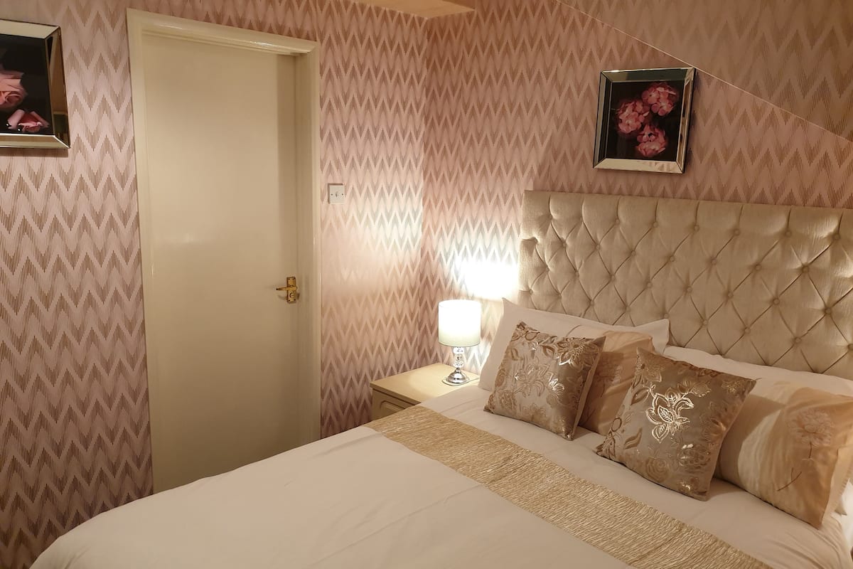 A bedroom with a tufted headboard, a bed with floral-patterned pillows, a bedside lamp, and a door with a patterned wallpaper.