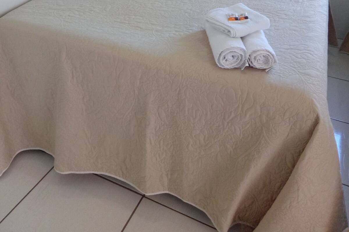 Un lit bien fait, avec un couvre-lit beige et des oreillers, une serviette pliée posée sur le lit et un petit objet décoratif sur l’oreiller.