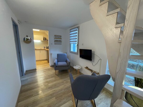 Ein modernes Wohnzimmer mit einem grauen Sessel, einem kleinen Tisch und einem an der Wand befestigten Fernseher. Im Hintergrund ist eine Küche mit einem Fenster und Jalousien zu sehen.