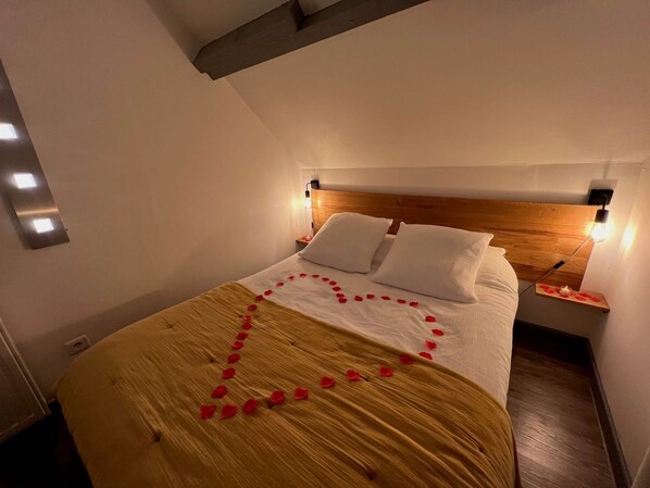 Una cama bien hecha adornada con pétalos de rosa formando un corazón, un cabecero de madera y lámparas de noche.