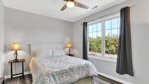 Ein Schlafzimmer mit einem Bett, zwei Nachttischen inklusive Lampen, einem Deckenventilator, einem Fenster mit Vorhängen und Blick ins Grüne.