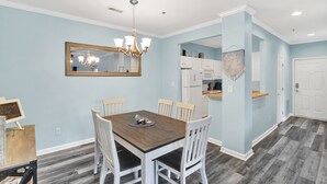Ein Essbereich mit einem Holztisch und weißen Stühlen, einer Kronleuchter und einer Küche im Hintergrund.