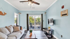 Ein Wohnzimmer mit Deckenventilator, einer Couch, einem Couchtisch, einem Fernseher und einer Glastür, die auf eine Terrasse im Freien führt.