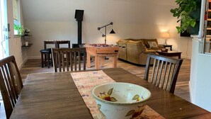 A dining area with a wooden table, chairs, a floral-patterned table runner, and a large bowl. There is a sofa, a lamp, and a plant in the background. - Country Getaway near Seattle (Maple Valley)