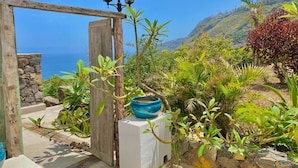 Um portão de madeira que dá para um terraço com vista para o mar, uma tigela azul sobre um pedestal e um muro de pedra.