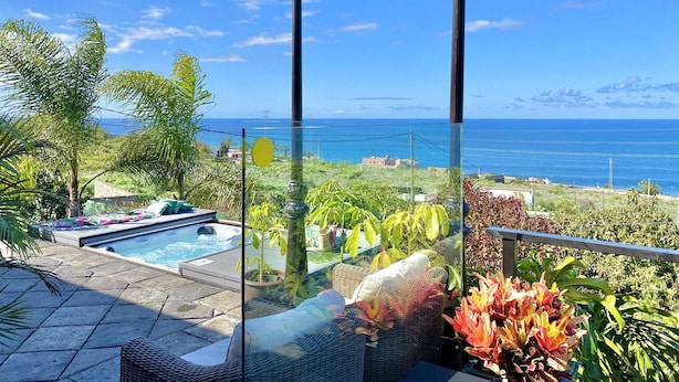 Área de piscina com guarda-corpo de vidro, mobiliário de vime e vista para o oceano.