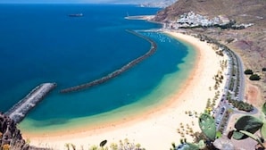 Paisagem costeira com uma praia curva, um cais e um povoado ao fundo.