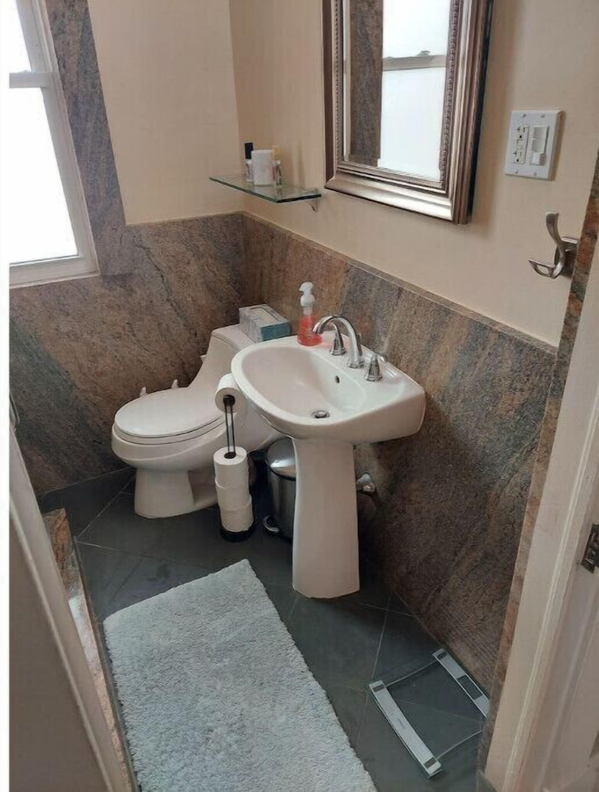 A bathroom with a toilet, sink, and mirror. There is a small shelf above the sink and a window to the left.