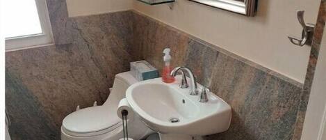 Une salle de bain avec des toilettes, un lavabo et un miroir. Il y a une petite étagère au-dessus du lavabo et une fenêtre sur la gauche.