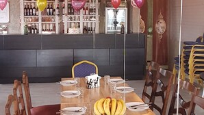 A dining area with a long wooden table set for a meal, surrounded by wooden chairs. There are balloons with names on them, a bowl of bananas, and a bar in the background with various bottles. - Green flower is located in the historical part of Kutaisi.5-minute walk  centre. (Kutaisi)