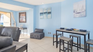 Una sala de estar moderna con un sofá gris, un pequeño reposapiés y un comedor con una mesa y taburetes. Hay obras de arte en las paredes y una ventana con vistas.
