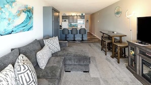 Un salon moderne avec un canapé gris, des coussins à motifs, une table basse et un téléviseur. On aperçoit une cuisine à l’arrière-plan, avec des tabourets de bar et une table à manger.