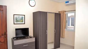 Um quarto com guarda-roupa, televisão sobre uma cômoda, quadro pendurado na parede e janela com cortinas.