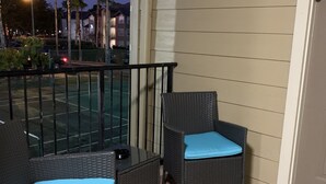 A balcony with two wicker chairs, a small table, and a view of a tennis court and palm trees.