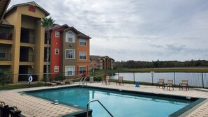A swimming pool with a tiled deck, surrounded by a fence, and adjacent to a multi-story building with balconies.