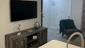 A living room with a flat-screen TV mounted on the wall, a wooden entertainment unit with a vase and a decorative item, a black sofa, and a chandelier.