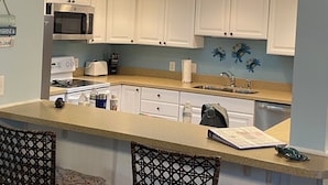 A modern kitchen with white cabinets, a central island, and a seating area with two chairs. - Beachwalk 106 Shipyard Hilton Head Island, 5 Min. Walk to Beach (Hilton Head Island)