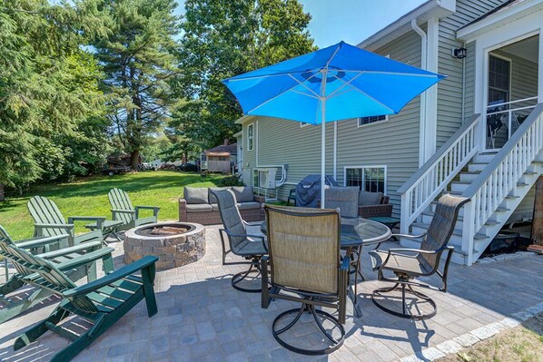 An outdoor patio with a fire pit, green Adirondack chairs, a round table, and a blue umbrella.