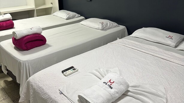 Habitación de hotel con dos camas, cada una con una colcha blanca y un almohadón con el logotipo del hotel. Hay una toalla blanca en cada cama, y un control remoto en la cama de la izquierda.