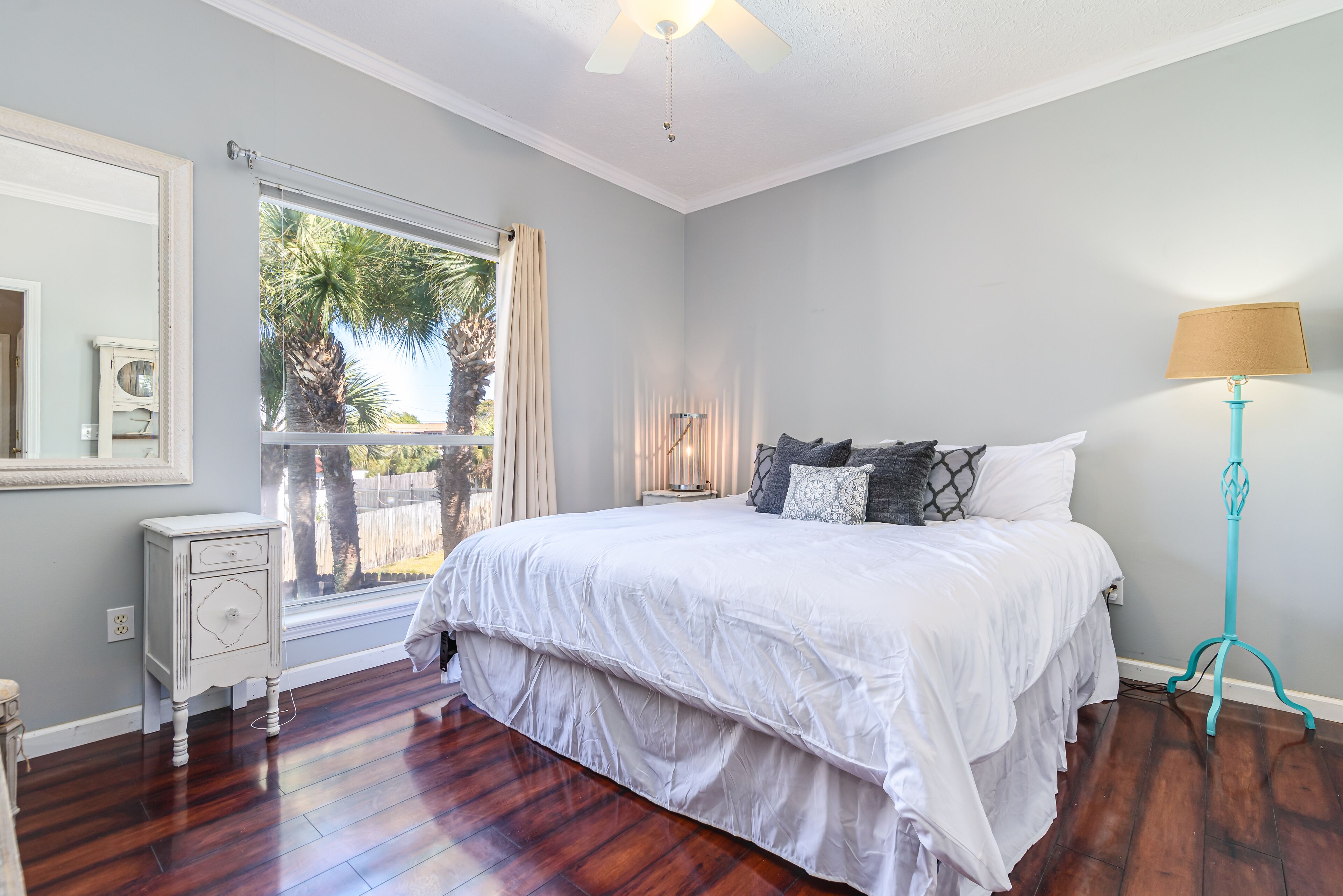 A bedroom with a large bed, a nightstand, a lamp, and a window with a view of palm trees.