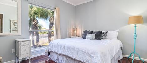 A bedroom with a large bed, a nightstand, a lamp, and a window with a view of palm trees.