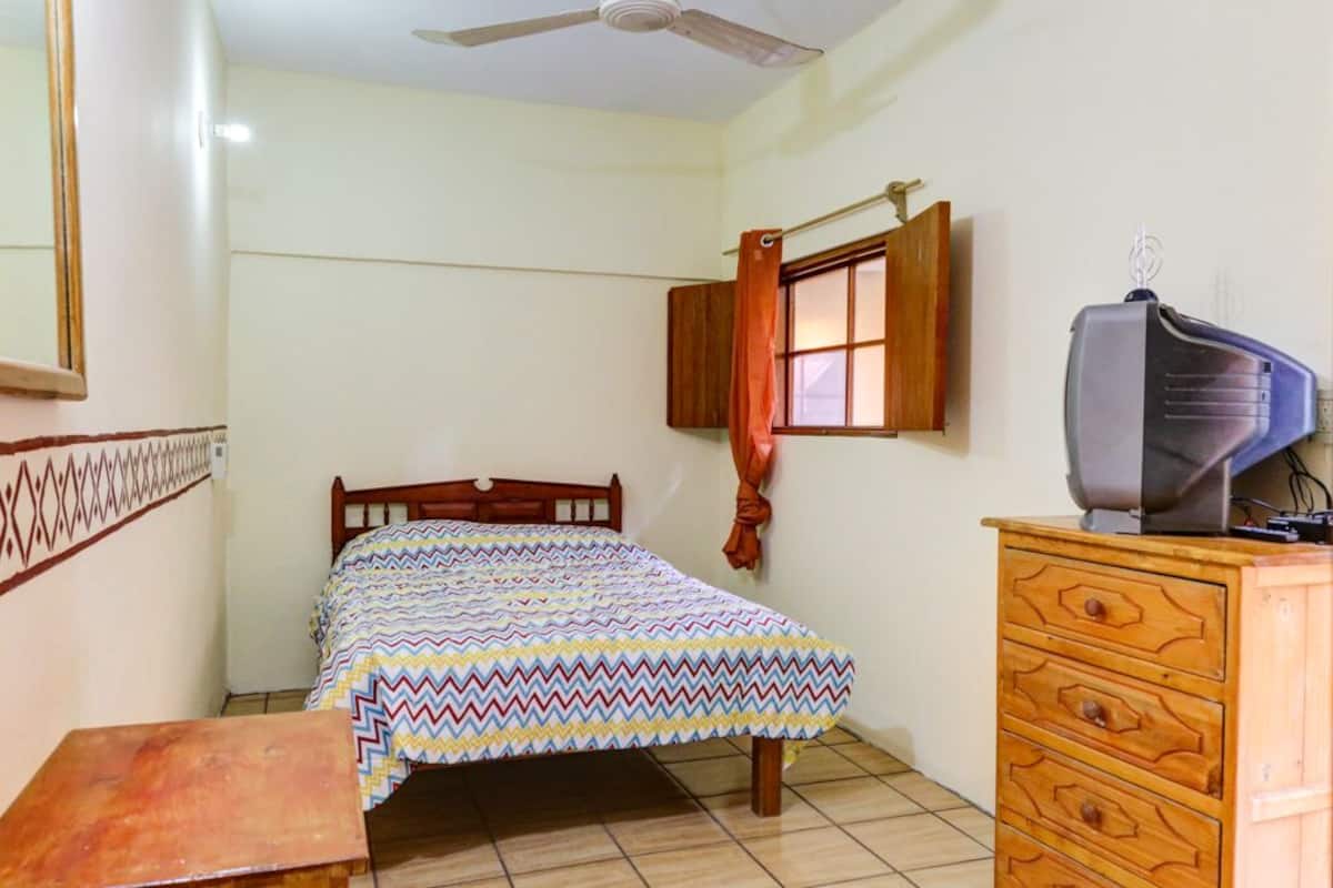 A bedroom with a bed, a wooden dresser, a television, and a window with curtains.