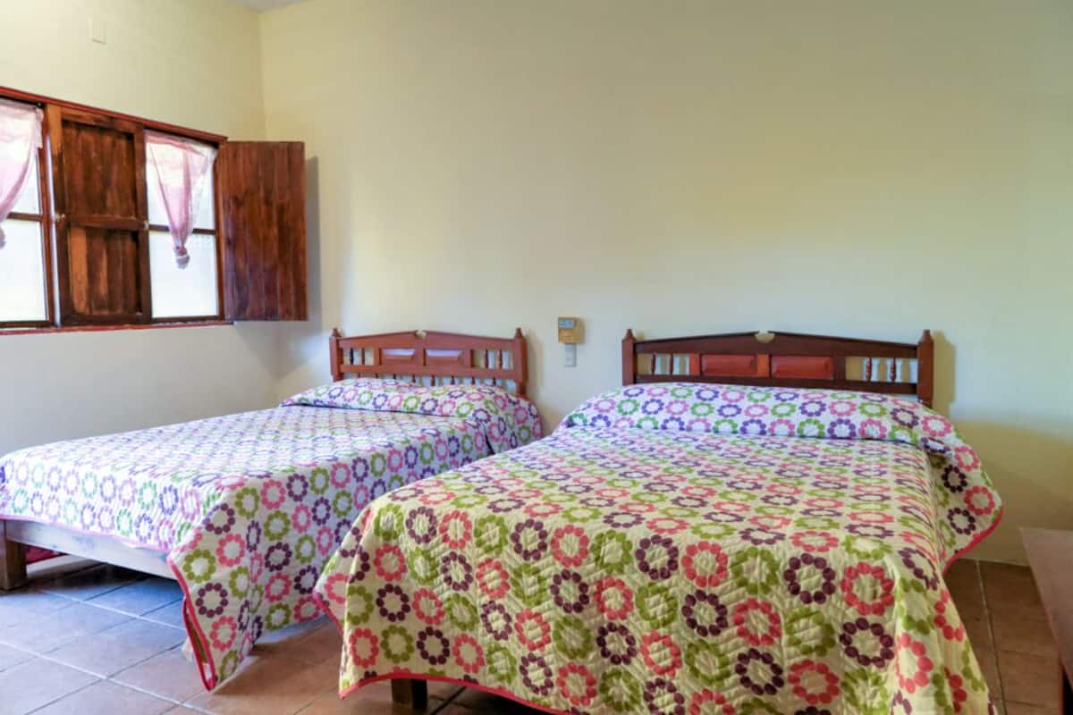A room with two single beds, each with a colorful patterned bedspread. There is a wooden headboard on each bed, and a window with wooden shutters on the left wall.