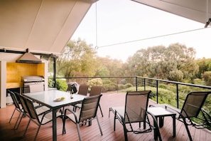 An outdoor patio with a grill, dining table, and chairs, overlooking a landscaped area with trees.