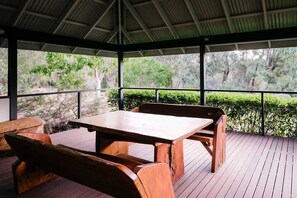 Un espacio de estar al aire libre techado con una mesa y bancos de madera, rodeado por una terraza de madera y vistas a la vegetación.