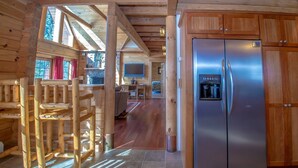 A log cabin interior with a wooden dining area, a kitchen with a stainless steel refrigerator, and a stone fireplace.