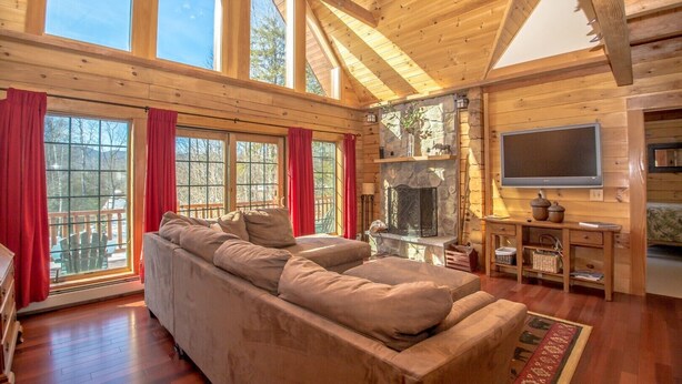 A log cabin interior with a large sofa, stone fireplace, and a TV mounted on the wall.