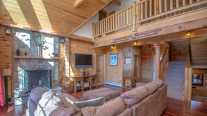 A log cabin interior with a living room, a stone fireplace, a television, and a staircase leading to an upper level.