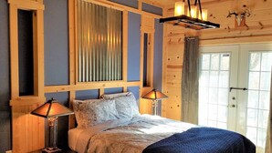 A bedroom with a bed, two bedside lamps, a wooden headboard, a window with curtains, and a door with a glass panel. - Blue Pine Lodge  - w/ Hot Tub - What a Mountain Cabin Should Be! (McGaheysville)