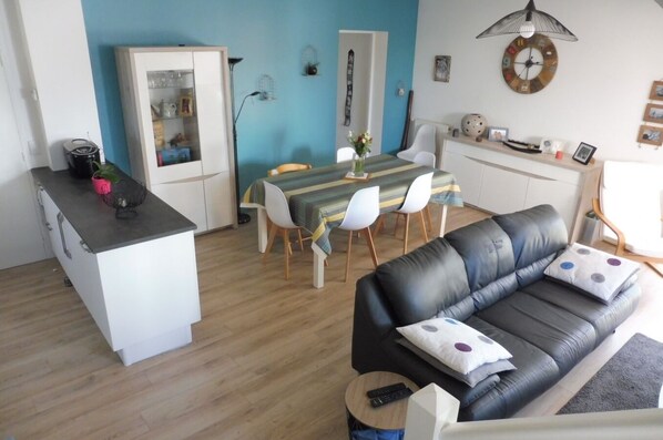 Un salon moderne avec un canapé en cuir noir, un coin repas avec une nappe à rayures et une cuisine avec des meubles blancs.